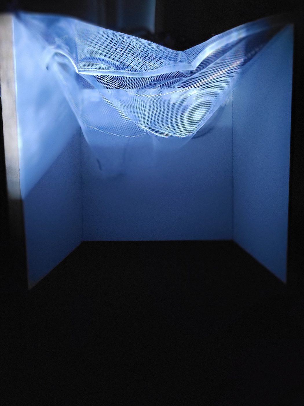 Scale model of the room in darkness: a sheet of fine mesh suspended across the upper corner, lit by a projection, glowing pale blue against white walls.